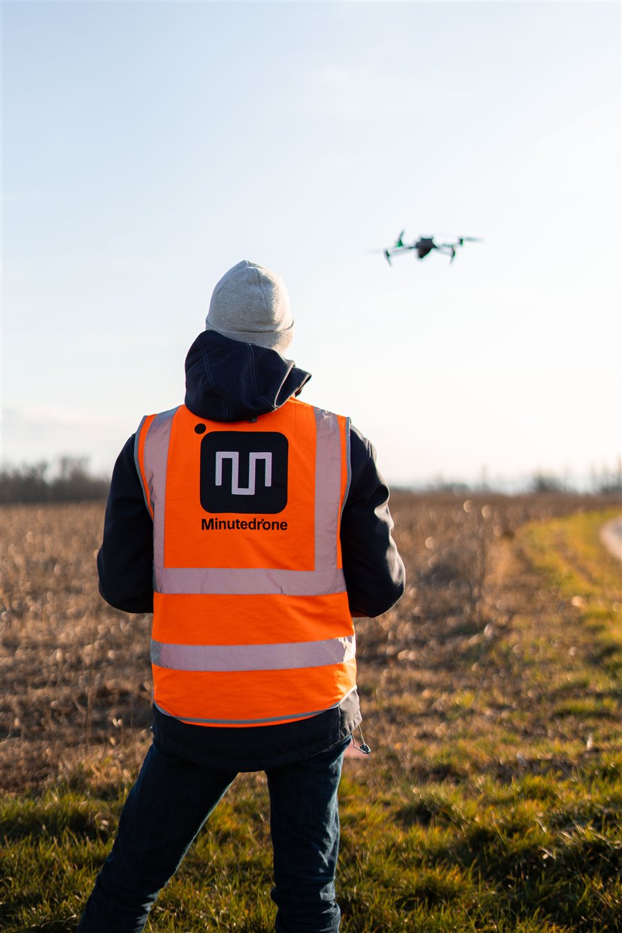 Télépilote de dos, en gilet orange Minutedrone, qui fait voler un drone au-dessus d'un champ.